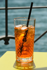 Close up view of a glass of Aperol Spritz on a table top with water in the background. No people.