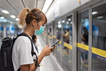 Asian woman with surgical face mask feel tired use smartphone standing waiting for subway, skytrain, carrying backpack, traveling to the city, social distancing, coronavirus, COVID19