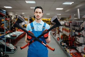 Male worker in uniform holds two axes, tool store