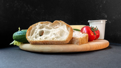 Two slices of white beautiful airy white bread, yogurt, fresh vegetables - cucumbers, tomatoes and celery, all on a round wooden cutting board on a black background close-up, side view.