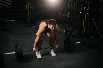 Obraz na płótnie Canvas Muscular strong man with perfect beautiful body wearing sportswear lifting heavy barbell from floor during sport workout training in modern dark gym. Concept of healthy lifestyle.