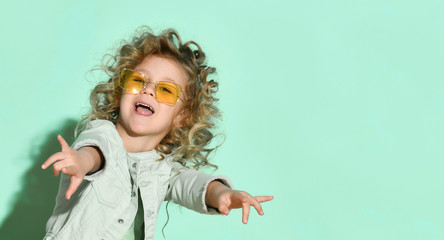 Nice little girl with curly hair in stylish glasses with orange lens laughing delightly stretching hands out. Close up portrait isolated on turquoise