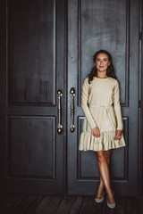 A beautiful young dark woman with dark curly hair and brown eyes in a gold dress is standing near the black door in the loft room. Soft selective focus.
