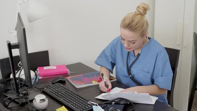 Young beautiful blond female doctor in blue coat, working at desk using computer and doing paperwork