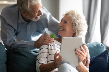 Happy grey-haired man embracing shoulders of pretty loving middle aged wife, communicating at home. Smiling mature woman using digital computer tablet, chatting talking with joyful husband indoors.