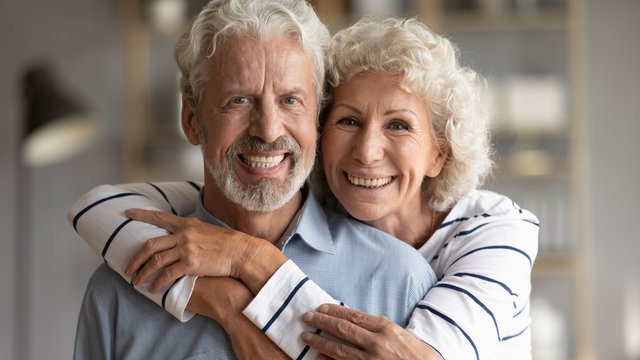 Head shot portrait of happy old retired family couple, enjoying sweet tender moment indoors. Loving affectionate middle aged hoary woman embracing smiling mature senior husband, looking at camera.