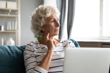Thoughtful mature senior woman sitting on couch with laptop on lap, looking away at window. Older...