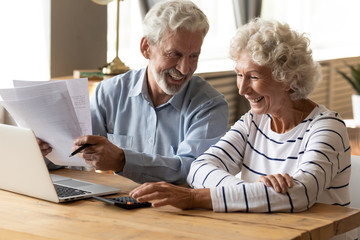 Happy middle aged family spouses sitting at table with computer and paper bills, calculating...