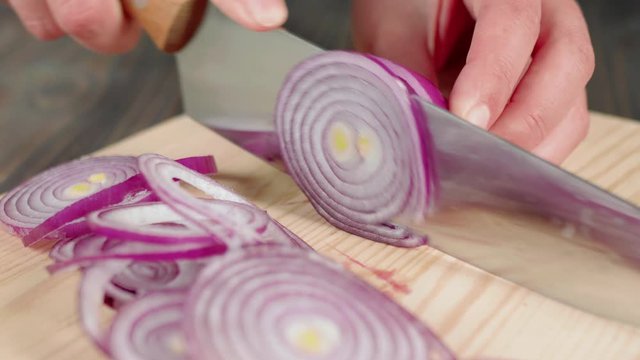 Woman slices with a sharp shiny knife a fresh purple onion on a wooden board. Concept of making a fresh summer salad from lettuce, cherry tomatoes, cucumbers and onions. Close up. 4K
