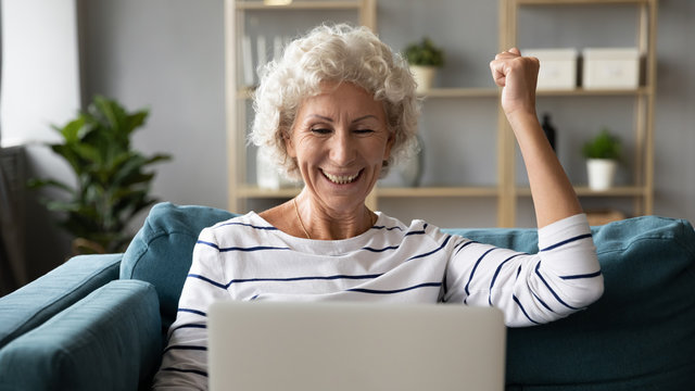 Front head shot view overjoyed elder senior woman reading online lottery win notification, celebrating success alone at home. Excited by good news middle aged grandmother laughing, making yes gesture.