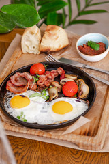 English breakfast. Fried eggs with fried sausages, bacon, tomatoes and mushrooms. On a wooden with bean paste.