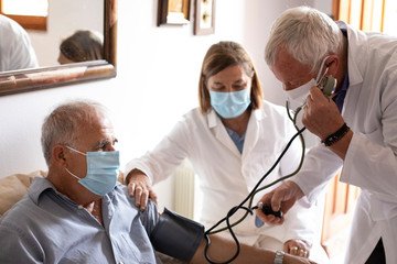 Anziano con mascherina viene visitato da un medico a casa che gli prende la pressione sanguigna...