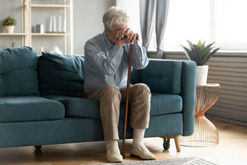 Full length exhausted disabled middle aged old man resting on sofa, leaning on walking cane,...