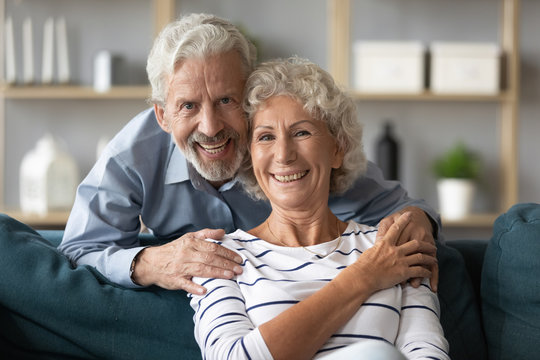 Head shot portrait of happy bearded senior elderly man cuddling smiling mature wife, resting on cozy sofa. Emotional positive retired married couple looking at camera, enjoying weekend time at home.