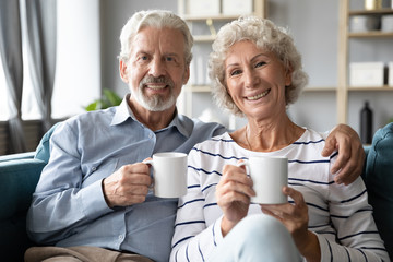 Portrait of happy middle aged married couple resting on comfortable sofa at home with cups of hot tea. Smiling retired family spouses enjoying lazy weekend morning time with coffee in living room.