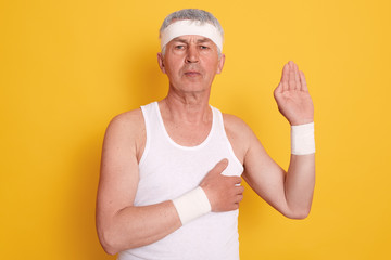 Studio shot of serious concentrated mature man posing against yellow wall, wearing white sleeveless t shirt and head band, showing gesture of honesty, patriotism and devotion, keeps hand on heart.