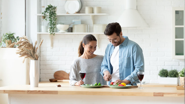 Happy millennial husband and wife stand in new kitchen prepare romantic dinner chop vegetables together, smiling young couple have fun cooking at home drinking wine celebrate anniversary