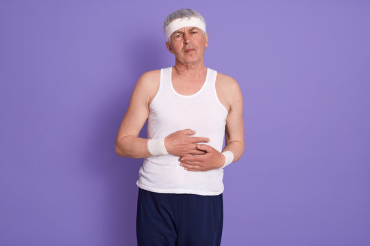 Picture Of Senior Man Dresses White T Shirt, Trousers And Head Band, Standing Against Lilac Studio Wall, Guy Has Abdominal Pain, Needs To Call Doctor Or First-aid. Health Care, People Concept.