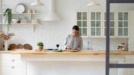 Young Caucasian woman stand at modern design kitchen counter cooking vegetable salad drinking wine,...