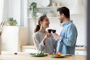 Obraz na płótnie Canvas Overjoyed young husband and wife cling glasses drink wine celebrate anniversary or moving at home kitchen, happy millennial couple tenants cheers cooking preparing food together in modern new kitchen