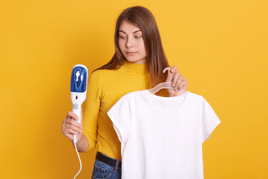 Closeup portrait of charming female with long hair, holding hangers with white casual t shirt and steam iron in hands, going on date with new boyfriend, posing isolated over yellow studio background.