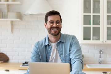 Happy millennial man in glasses sit at the table in modern kitchen talk laughing, smiling young...