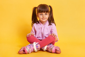 Closeup portrait of calm little girl sitting on floor with crossed legs, wearing casual attire and rolling skates, having dark hair and two ponytails, looks at camera, keeping palms on knees, has rest