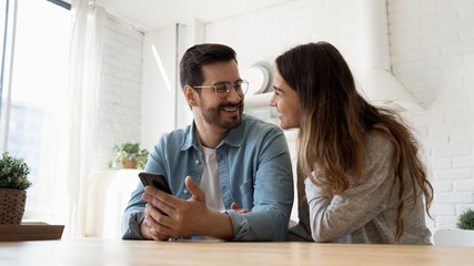 Overjoyed husband and wife sit at table in modern design kitchen laugh enjoy family weekend using modern smartphone gadget together, happy couple relax at home have fun browsing Internet on cell