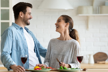 Happy millennial couple laugh enjoy meal drink wine in modern new kitchen, overjoyed young husband and wife cook prepare food at home, enjoy romantic domestic date, celebrate anniversary together