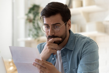 Frustrated millennial guy in glasses read bad unpleasant news in postal letter, sad confused young...