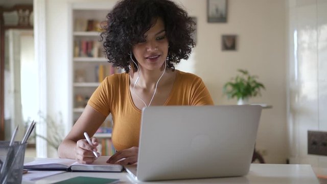 Young woman student using laptop to study online from home in internet Spbd. millennial girl writing in notebook looking at screen listen to teatcher and nod. headphones. distance learning quarantine