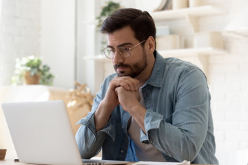 Pensive young Caucasian man in glasses sit at desk look at laptop screen thinking pondering,...