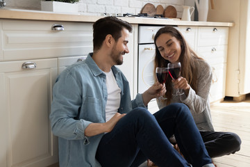 Overjoyed millennial couple sit on wooden floor in modern design kitchen drink wine celebrate...