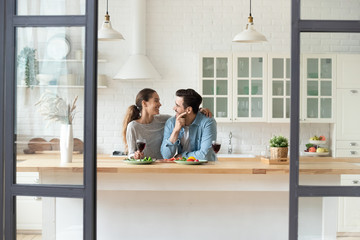 Happy Caucasian millennial husband and wife relax drink wine in modern new home kitchen, smiling...