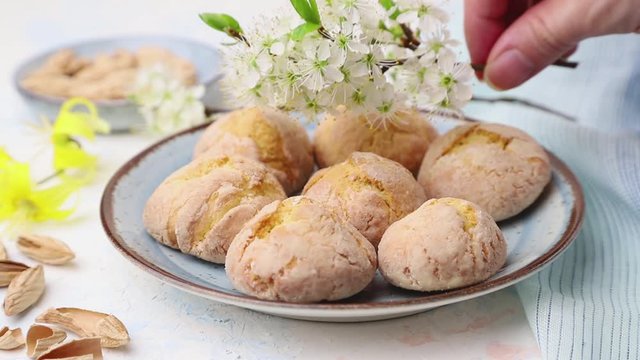 male hand decorate almond cookies with cherry flowers on ceramic plate on a white background with blue textile. Side view, close up.