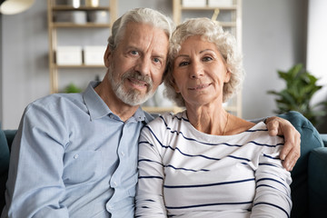 Portrait of affectionate mature retired married couple cuddling embracing on comfortable sofa. Loving caring middle aged husband hugging hoary senior wife, looking at camera, good relations concept.