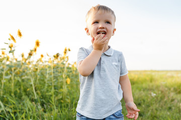 Little boy playing on the background of countryside and smiling. Three years kid having fun in field of sunflowers on sunset. Summer outdoors lifestyle, child leisure, childhood