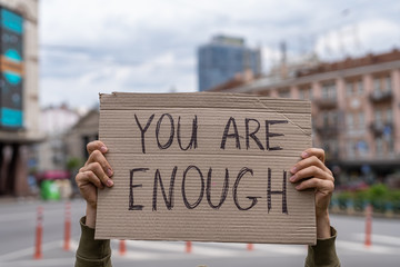You are enough support phrase for body positive movement. self-respect and self love support.  Hands holding banner outside on city streets. social care motivation concept. Friendly mood attitude.