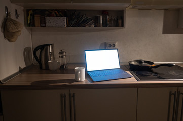A working laptop with a white screen stands on the kitchen table next to the stove in the dark. The concept of remote work. Quarantine during coronavirus.