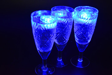 Crystal champagne glasses with carvings in the form of snowflakes are located on a black matte background. Bright multicolor doidic glowing ice cubes float in the drink.