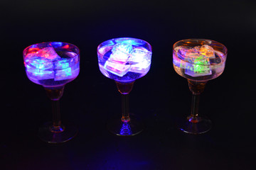 Three open champagne glasses with luminous multicolor ice cubes are located on a black matte background.