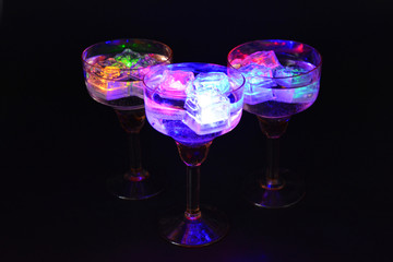 Three open champagne glasses with luminous multicolor ice cubes are located on a black matte background.