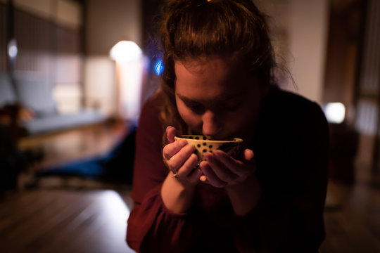 Young woman tourist seiza sitting in traditional Japanese restaurant dark night room in ryokan, holding cup of green sencha tea drinking