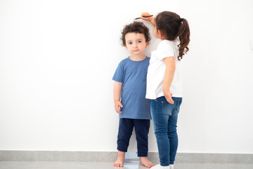 Cute girl measuring height of a funny child with curly hair. Sister measures the growth of her brother at a blank white wall.