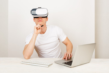 Male using virtual reality headset interacts with augmented things orienting in three dimensional space while sitting on his desk with keyboard and laptop on white background.