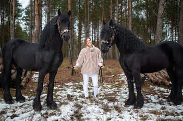 Handsome caucasian woman with fair hair in warm clothes goes for a walk with two black horses in winter forest.