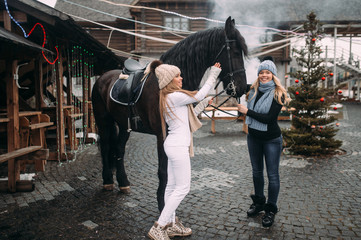 Picture of two young cheerful female riders in warm jackets, trousers, boots, scarfs and knitted hats with a horse outside