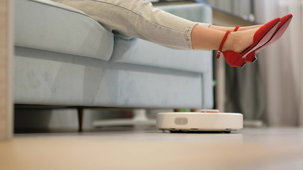 Robotic vacuum cleaner cleaning the room while woman resting on sofa.