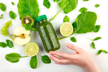 A woman's hand holds at her fingertips a bottle of green detox, wich is surrounded by fresh fruits,...