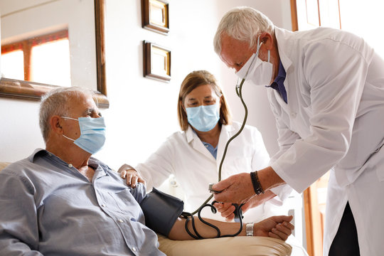 Anziano con mascherina viene visitato da un medico a casa che gli prende la pressione sanguigna insieme ad un altra dottoressa che assiste